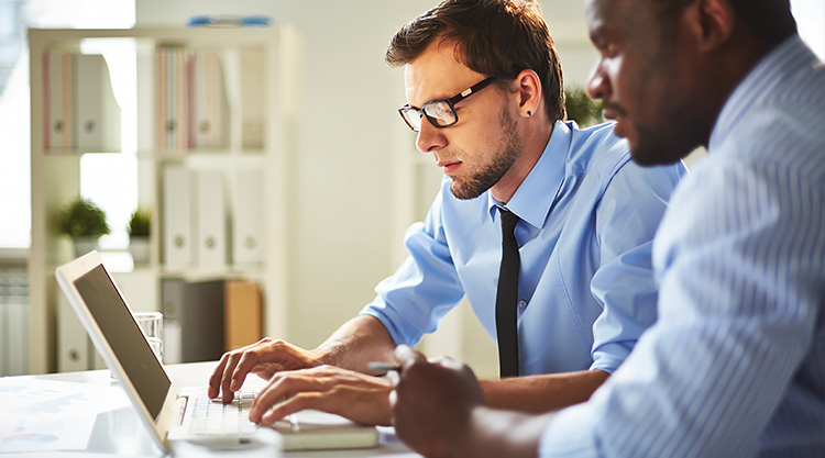 A man working on his laptop and a coworker seated next to him looking over