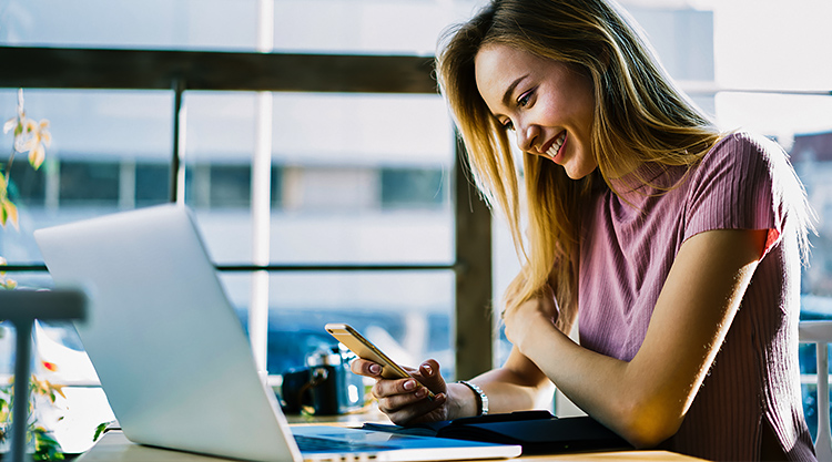 A smiling young woman looking down at her cell phone with her laptop open in front of her while seated in a restaurant