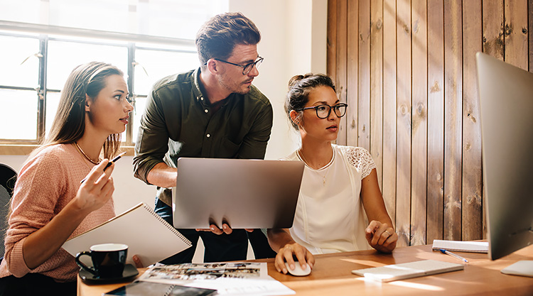 A group of coworkers seated at a desk and looking at their computer monitor