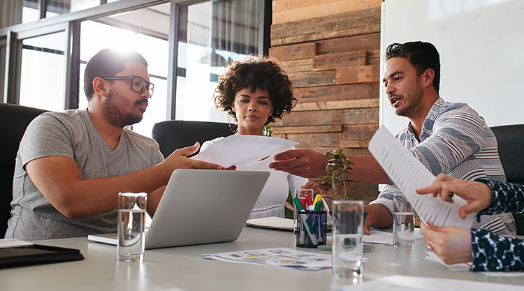 Three people are seated at a conference table and sharing files with each other in an office space