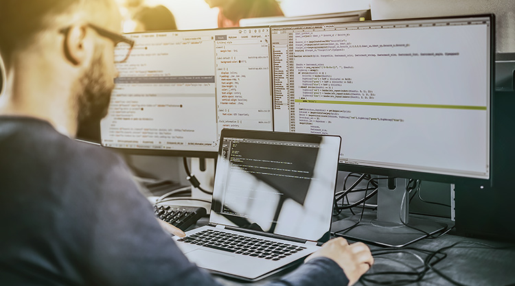 An image of a man seated in front of two computer monitors and a laptop