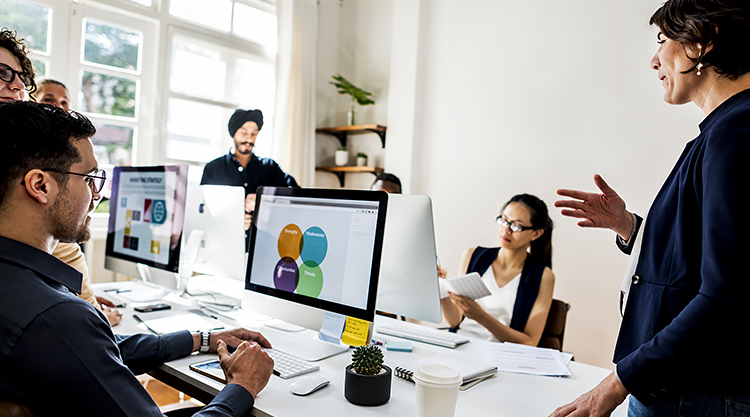 A group of co-workers typing on their computers with one woman standing in front of them