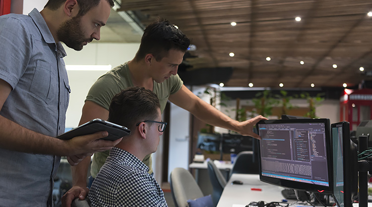 Three men stand near a computer monitor looking at the screen intensely in an office space