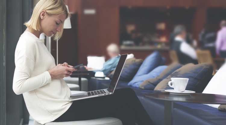 A woman texting on her phone with her laptop on her lap in a restaurant