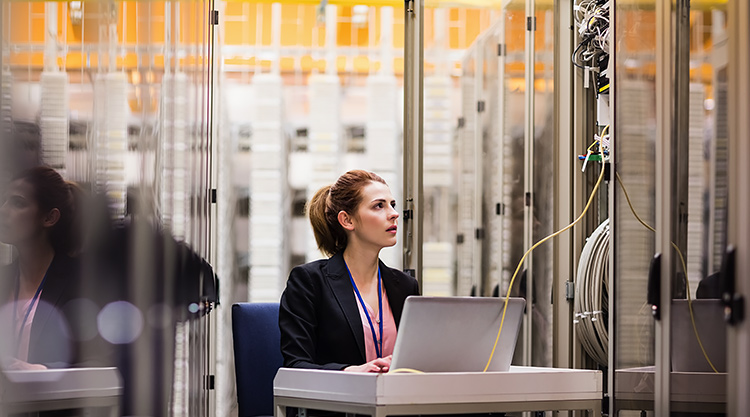 A woman is seated at a desk staring at a wire connected to the laptop in front of her