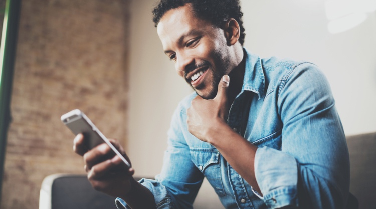 A smiling man looking at his cell phone while seated on a couch