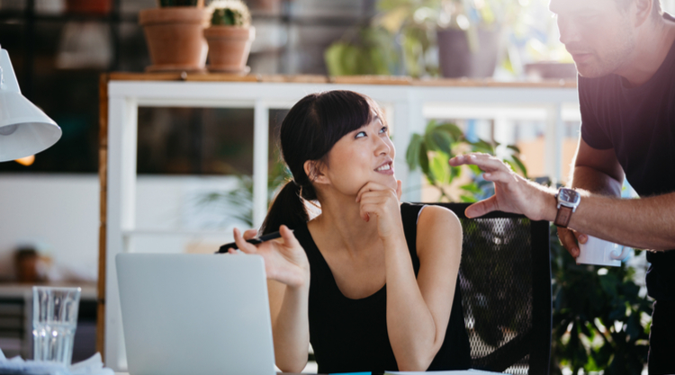 A smiling woman looking up at the man next to her while working on her laptop in an office