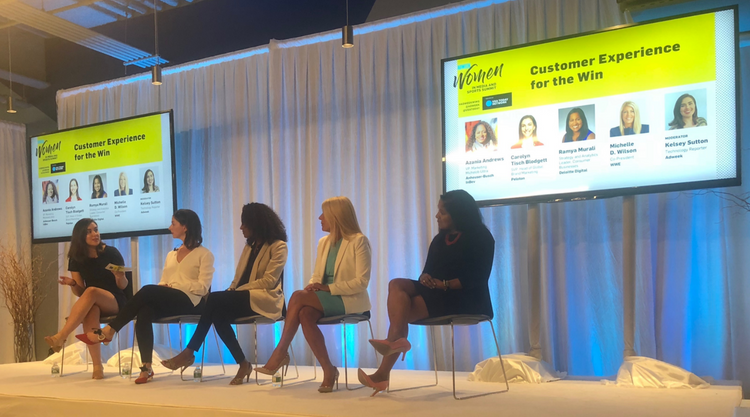 An adult woman sitting on stage and speaking to the four other adult women seated next to her and listening. Behind them are two monitors with the text 