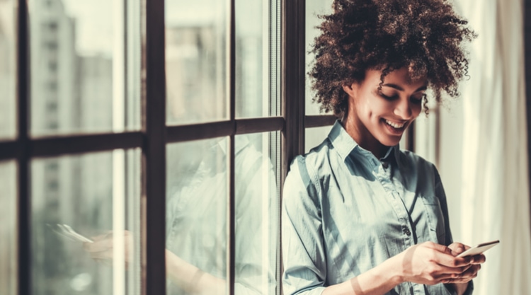 A smiling woman looking at her cell phone and leaning against a window