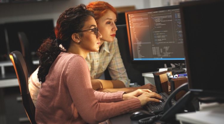 A young woman typing on her computer keyboard while another woman leans over to watch