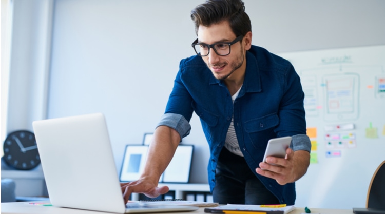A man holding a cell phone while working on his laptop in an office