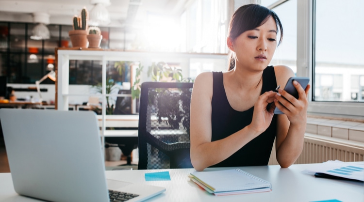 A woman looking at her cell phone with her laptop open in front of her while working in a well-lit office space