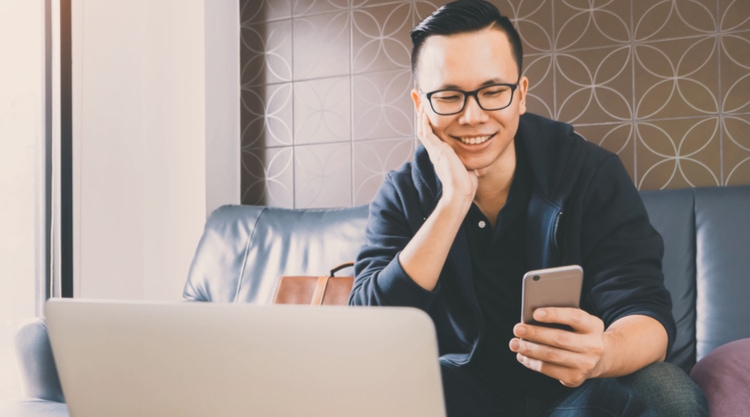 A smiling young man holding a cell phone and working on his laptop while sitting on a couch in an office