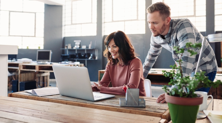 A smiling woman typing on a laptop in an office with a man leaning over and looking at the screen