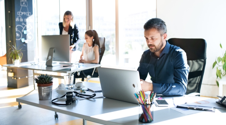 A man working in an office space at his desk with two women seated behind him