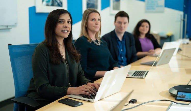 Four adults dressed in business work seated in a conference hall with their laptops open