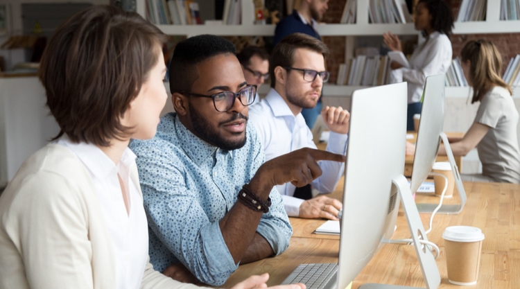 An adult man is pointing at their coworkers computer screen and they are both in an office filled with workers