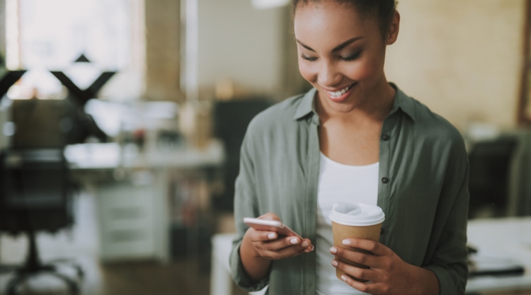 A young woman looking at her cell phone and holding a mug in her hand while standing in an office