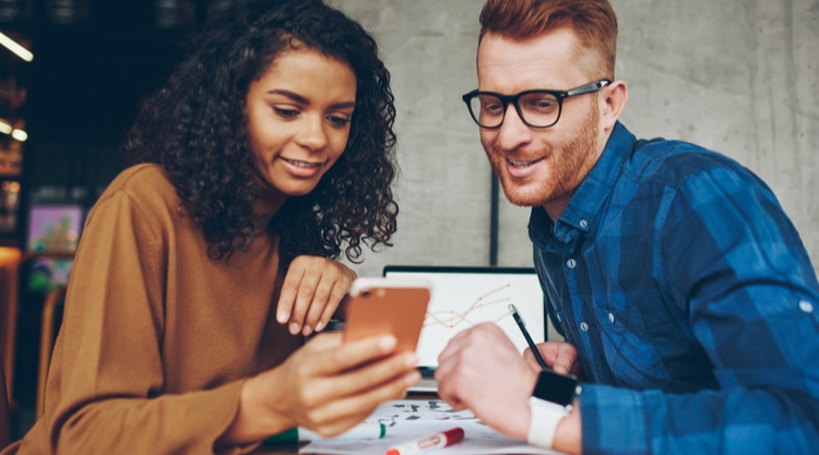A woman holding a cell phone and sharing that screen with the man next to her in an office