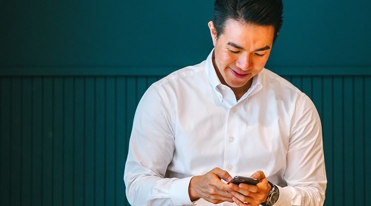 An adult man typing on his phone while standing in front of a blue-green wall