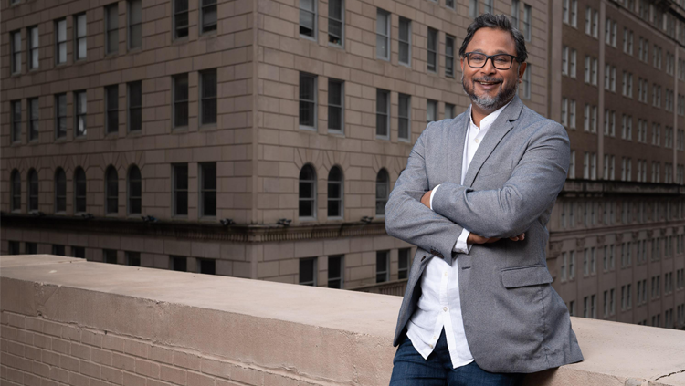 Image of a smiling adult man leaning against a building with a backdrop of a city behind him