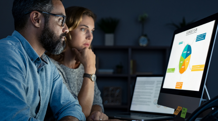 A man and woman looking at a computer screen with a pie chart displayed in a dark room