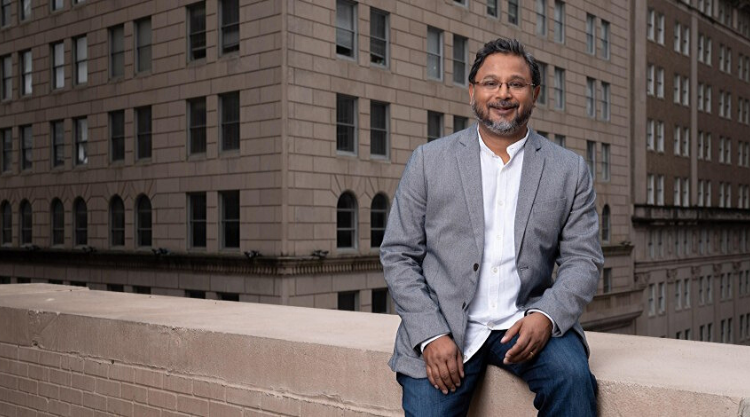 A photo of a man leaning on a building ledge with a backdrop of a city behind him