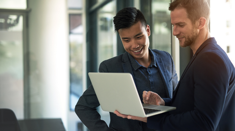 A smiling young man looking at a laptop screen being held up by the man standing next to him