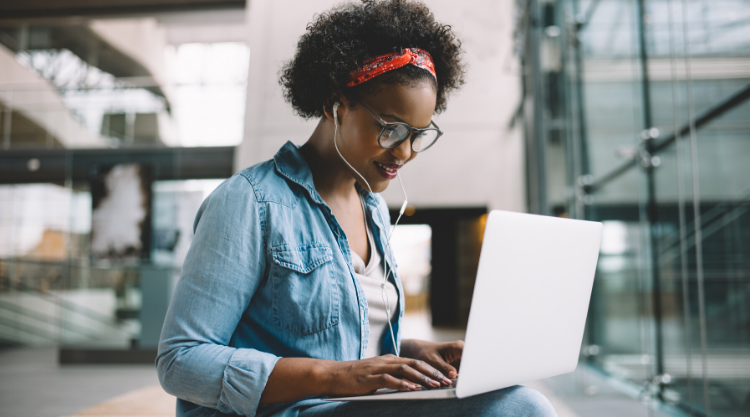 A woman with head buds in typing on her laptop while sitting outside an office