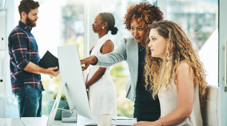 A businesswoman standing in an office points at a computer screen while a woman seated follows along. There are two other employees behind her