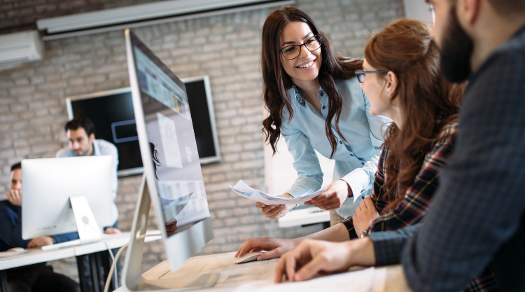 A smiling woman carrying documents in an office walks ups to two seated coworkers.