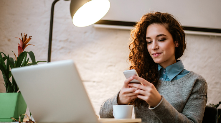 A young woman holding a cell phone is seated in a cafe with her laptop open in front of her and a cup of coffee.