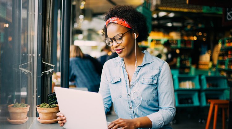 A woman seated in a cafe with an earbud in her ear and looking at her laptop.