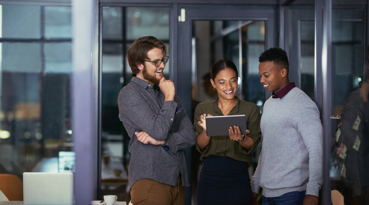 A woman stands next to two men holding a tablet in the corner of an office