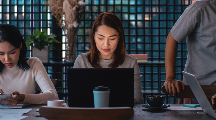 An adult woman working on her laptop in a cafe with a man and woman next to her.