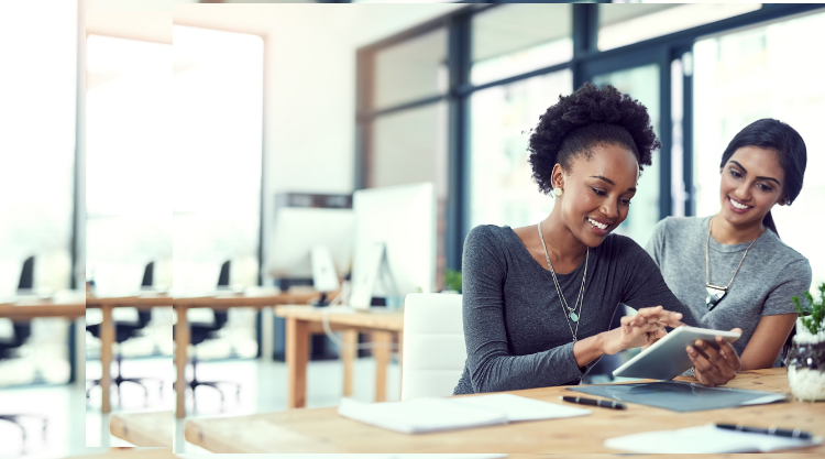 A woman holds a tablet with another woman leaning over and looking at the screen. They are both in an empty office