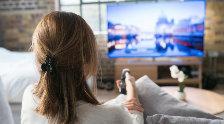 A young woman sits on her couch while holding a remote. In front of her is a blurred-out room and television.