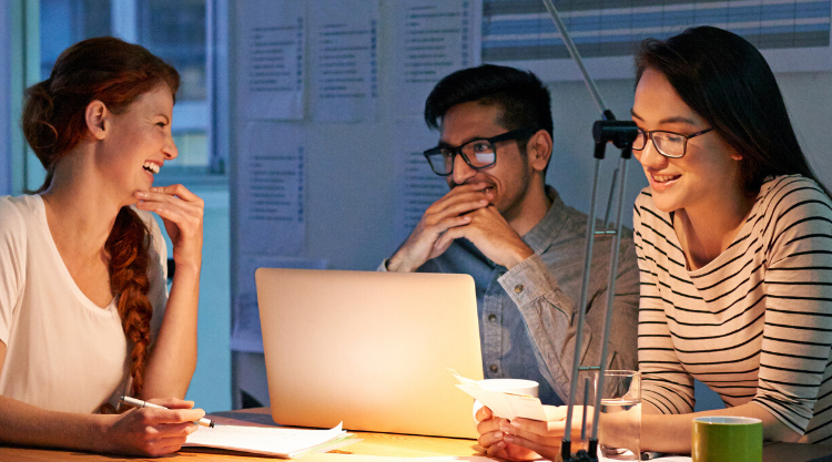 Three laughing adults in an office work together late into the day