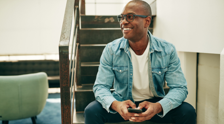A smiling adult man sits at the bottom of a staircase using his cell phone.