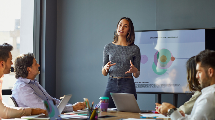 A woman is standing in the center of a conference room giving a presentation with a crowd of people listening.