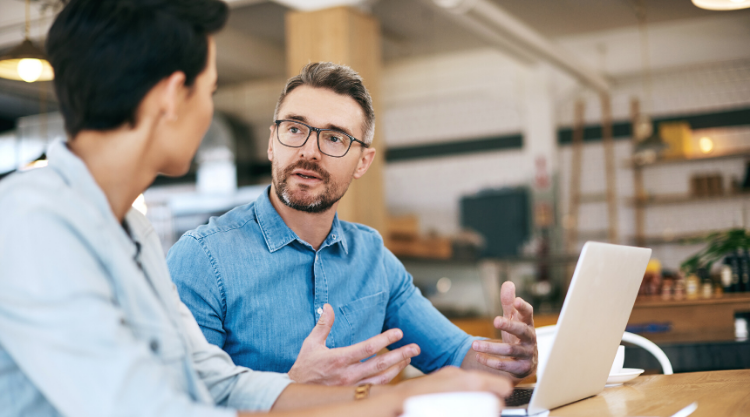 A man is talking to his associate who's seated next to him working on his laptop in a cafe.