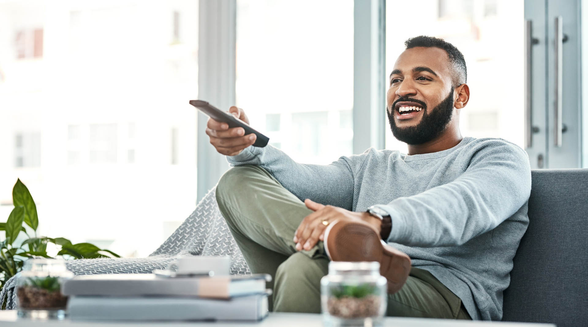 A smiling adult man holding a remote control seated on a couch in his home