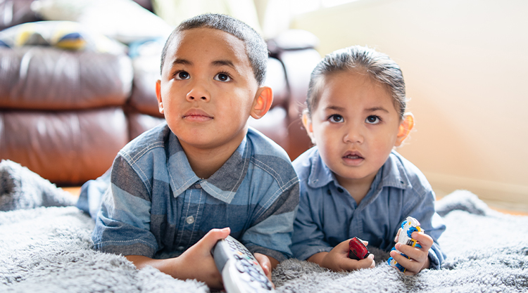 A small toddler and child lay on blanket while holding a remote control