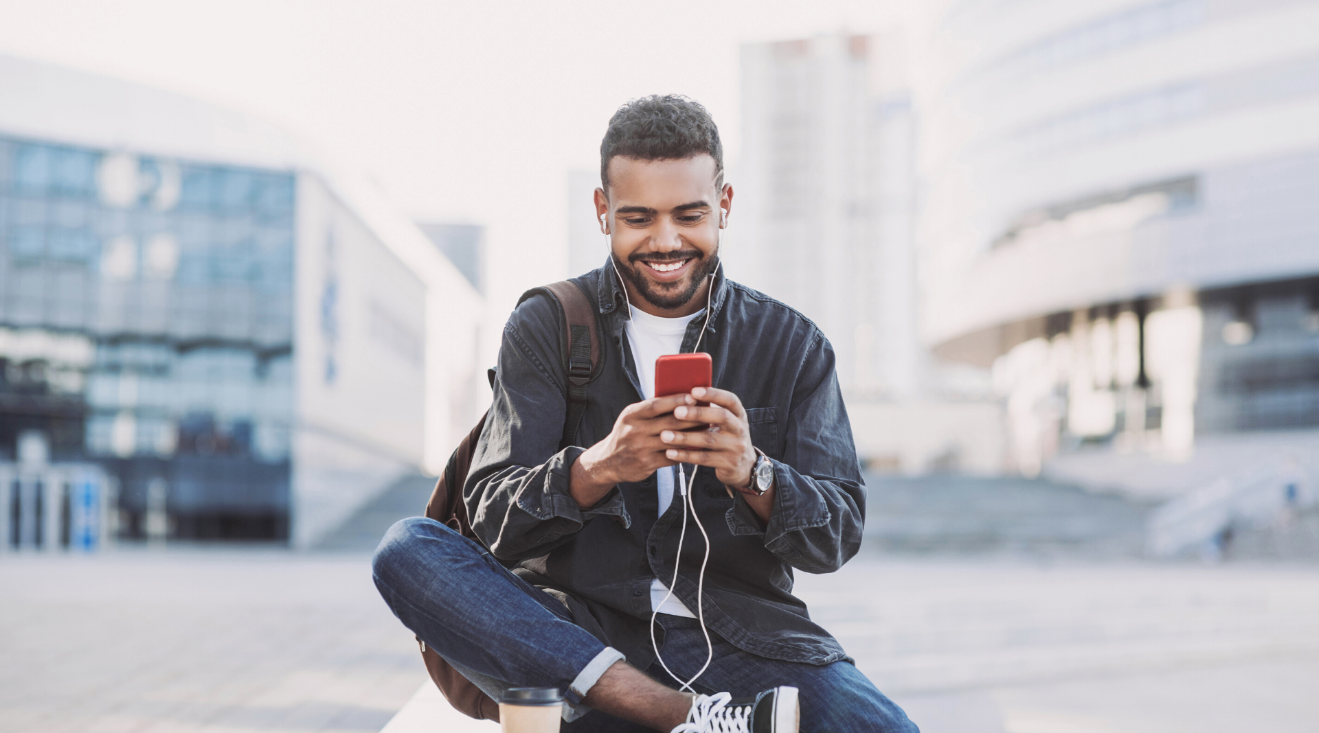 A young man looks into his cell phone while seated on a bench in the center of a public area