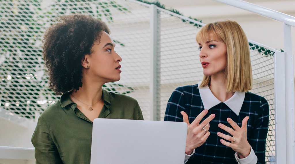 Two women sit next to each other talking while working outside.
