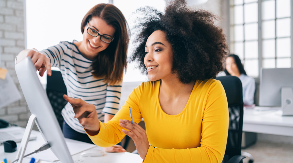 A woman sitting at her desk points at the computer screen while the woman next to her looks over