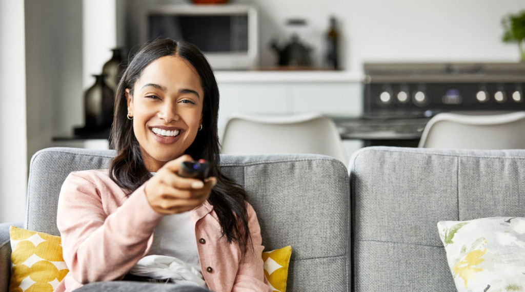 A smiling young woman seated on her couch holding a tv remote.