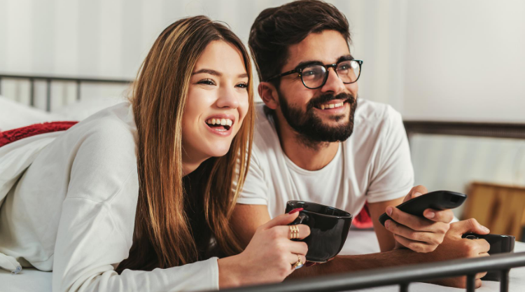 A smiling couple laying in bed while holding a mug and TV remote.