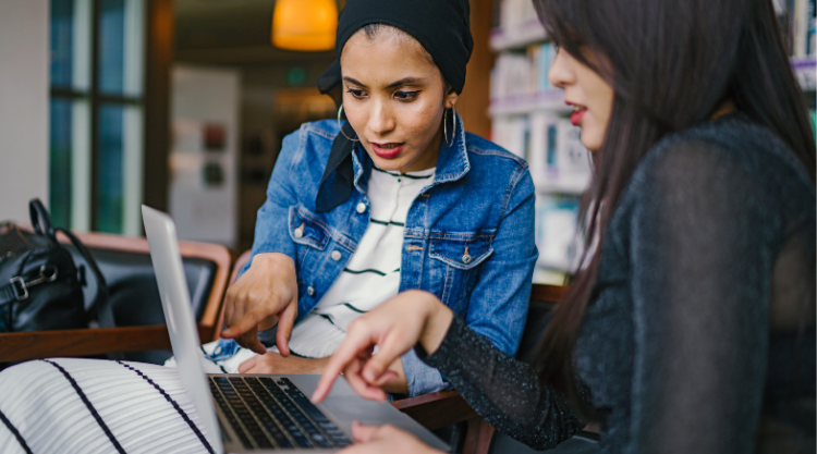 Two young women in a library working together and looking at the laptop screen in front of them.