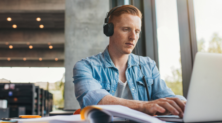 A man is working on his laptop with headphones while in a library.